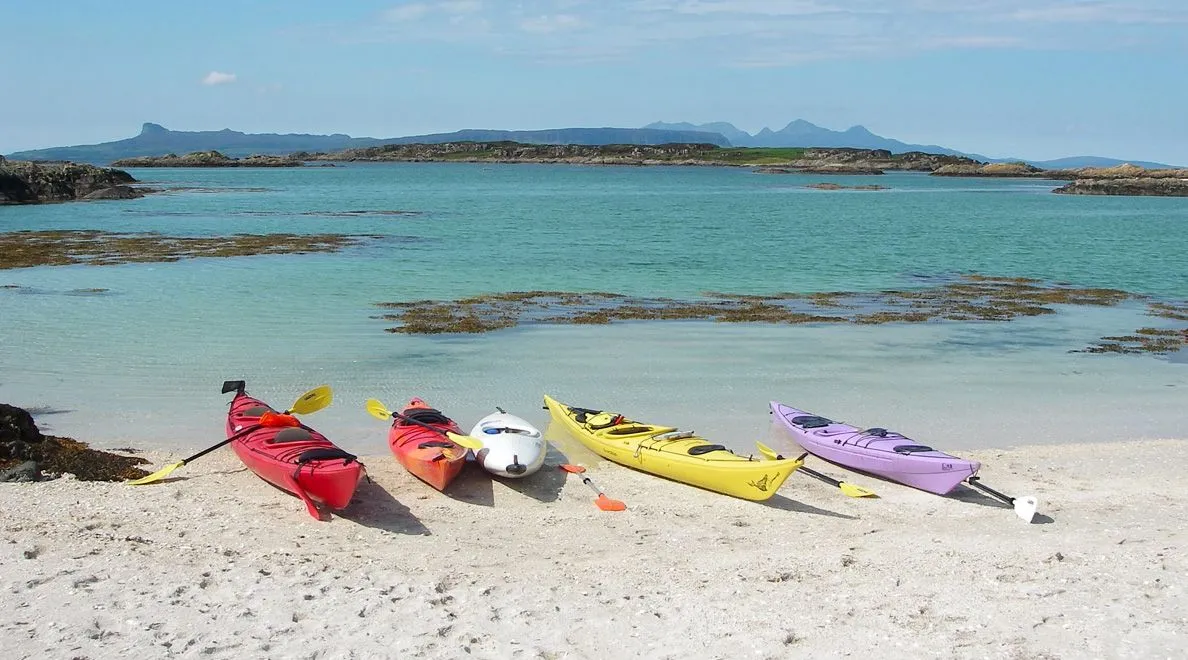 A vibrant outdoor image depicting several colorful kayaks resting on a sandy beach beside clear, turquoise water under a blue sky with distant landmasses.