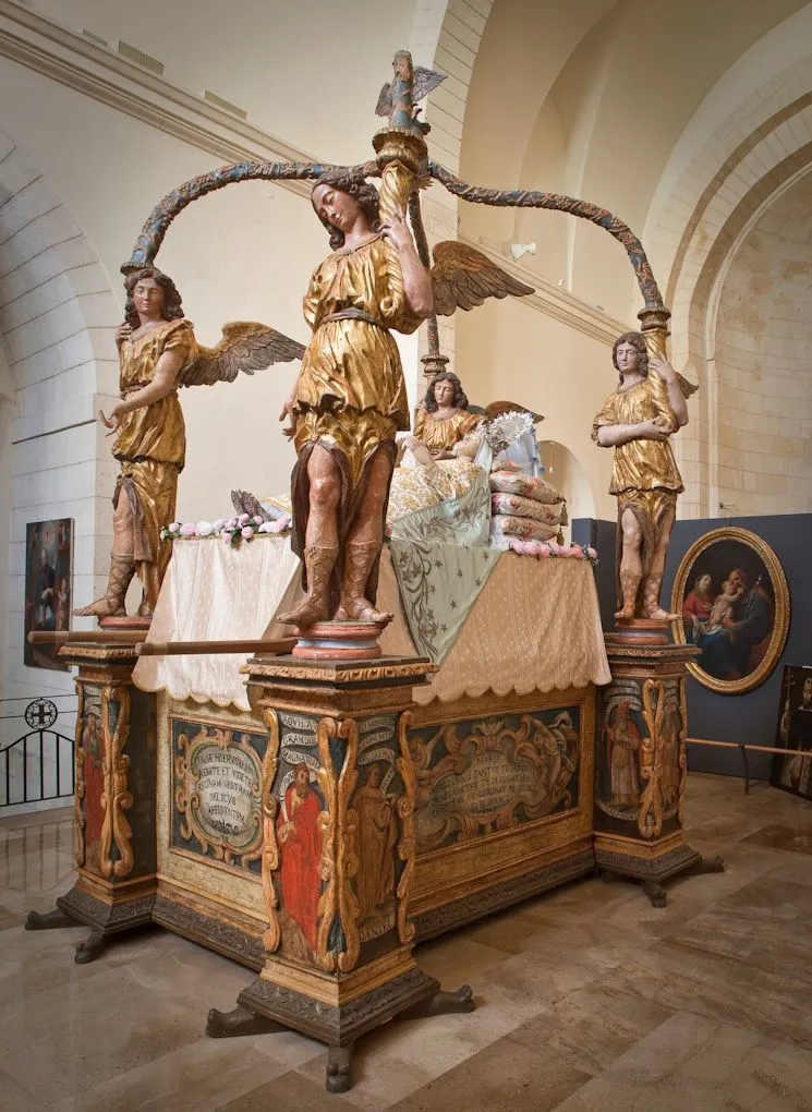 The image features a large, ornate wooden structure adorned with multiple golden statues of angels, likely an ancient altar or canopy, prominently displayed within what appears to be a museum or church setting.