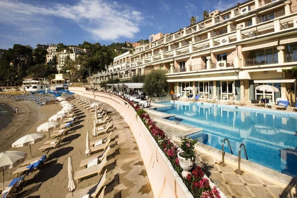 A luxurious multi-story hotel or resort building with a large outdoor swimming pool and a private beach area lined with lounge chairs and umbrellas, set against a backdrop of hills under a clear blue sky.
