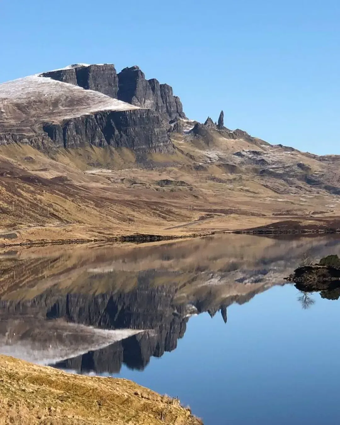 A picturesque landscape photo featuring the Old Man of Storr and surrounding mountains reflected in a calm loch under a clear blue sky.