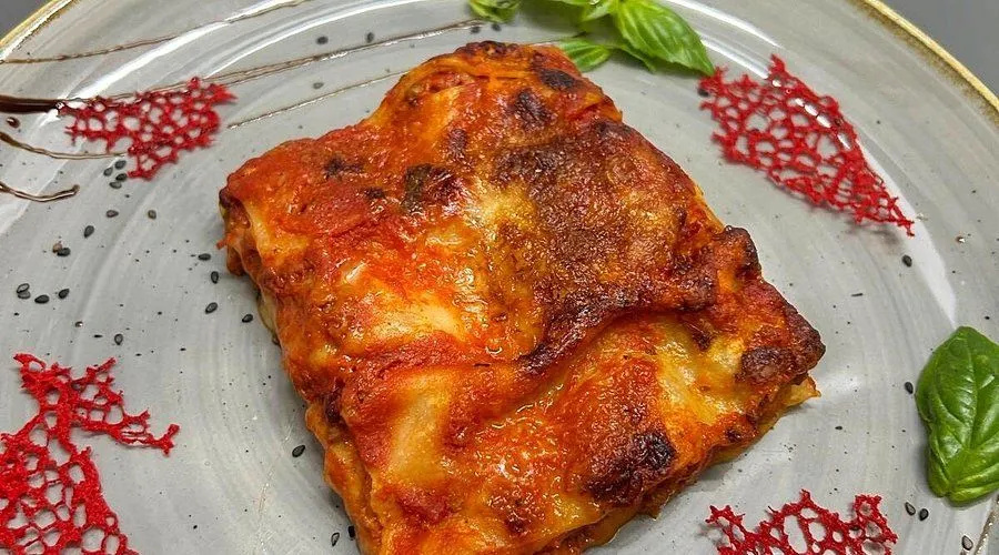 A close-up shot of a baked lasagna portion served on a plate, garnished with basil leaves and a small decorative element. The lasagna has a rich, golden-brown crust and visible layers of pasta, cheese, and tomato sauce.