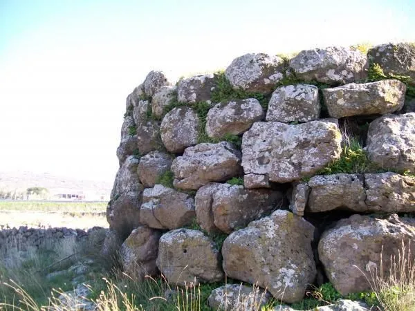 An image capturing a section of an ancient-looking stone wall, likely part of a historical or natural structure, built from large, irregular stones with moss and grass growing between them. The wall is seen from a low angle against a bright sky, with some sparse vegetation in the foreground and a distant, blurred landscape.
