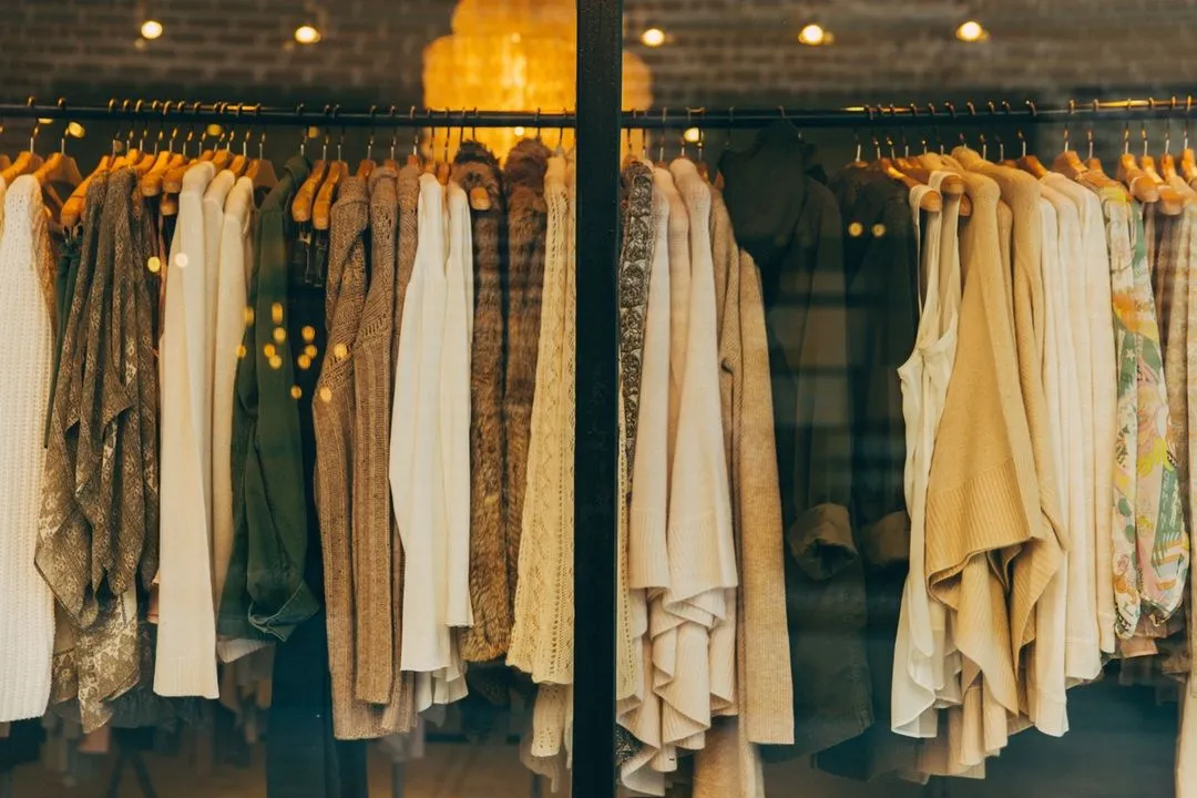 An editorial style image showcasing a variety of clothing items on hangers within what appears to be a retail store or boutique window. The focus is on the textures and styles of the garments.