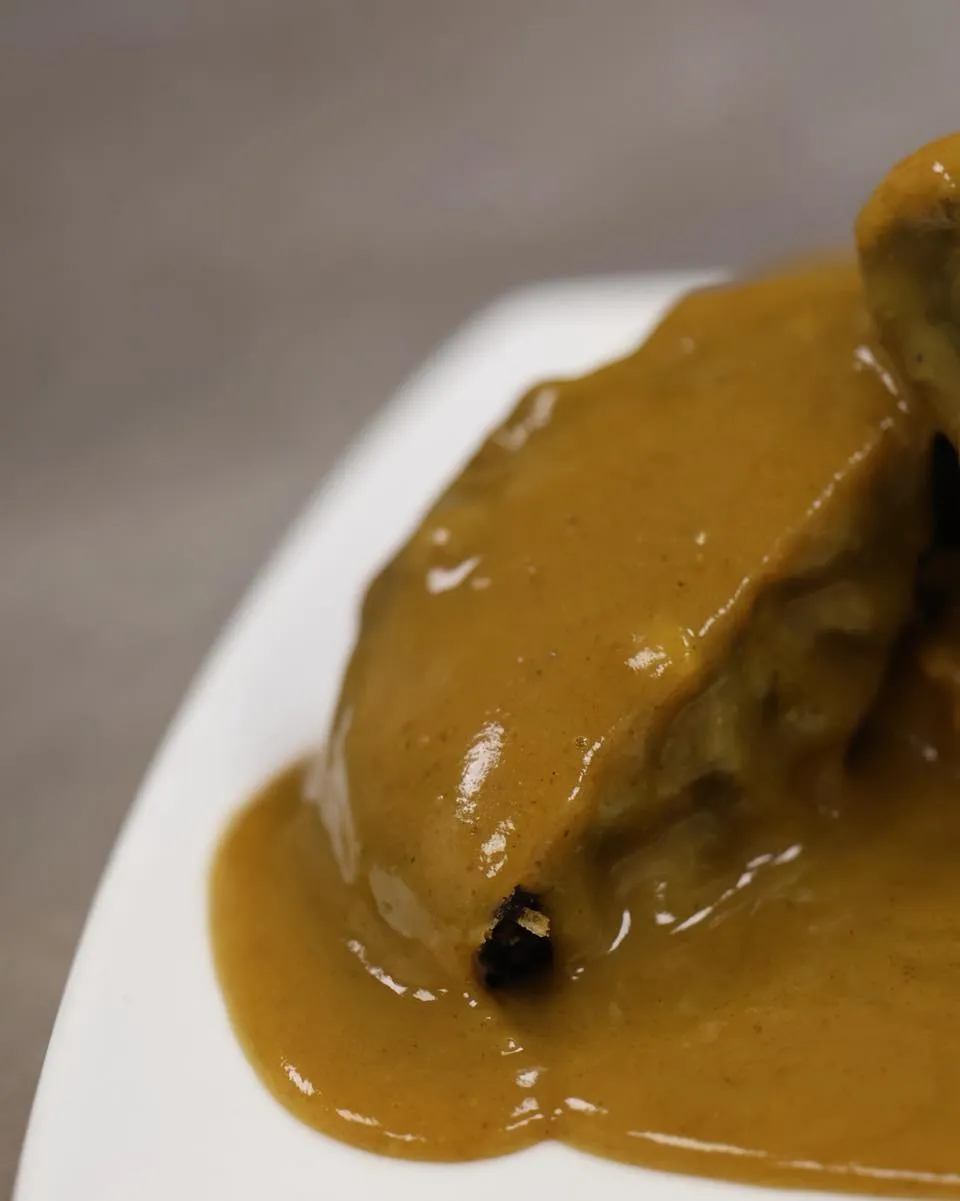 A close-up, professional photograph of a food item covered in a rich, golden-brown sauce, served on a white plate. The image is sharp, well-lit, and focuses clearly on the textured food and glistening sauce, with a softly blurred background. The colors appear natural and appealing, enhancing the food's realism.