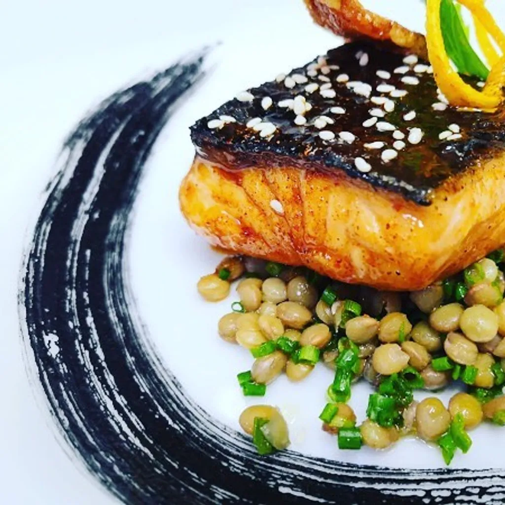 A well-composed and professionally taken close-up of a salmon dish with lentils and an artistic plate design. The lighting is excellent, highlighting the textures and vibrant colors of the food. The main elements, the salmon and lentils, are sharp and in focus, contributing to the overall photo-realistic quality. The colors appear accurate, making the dish look appetizing.