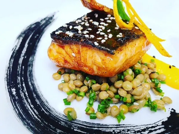 A well-composed and professionally taken close-up of a salmon dish with lentils and an artistic plate design. The lighting is excellent, highlighting the textures and vibrant colors of the food. The main elements, the salmon and lentils, are sharp and in focus, contributing to the overall photo-realistic quality. The colors appear accurate, making the dish look appetizing.