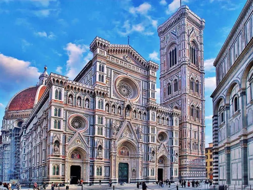 A vibrant, well-composed image of Florence Cathedral (Duomo di Firenze) and Giotto's Bell Tower under a bright blue sky. The intricate details of the marble facade are clearly visible, with some people visible in the piazza in front of the building.