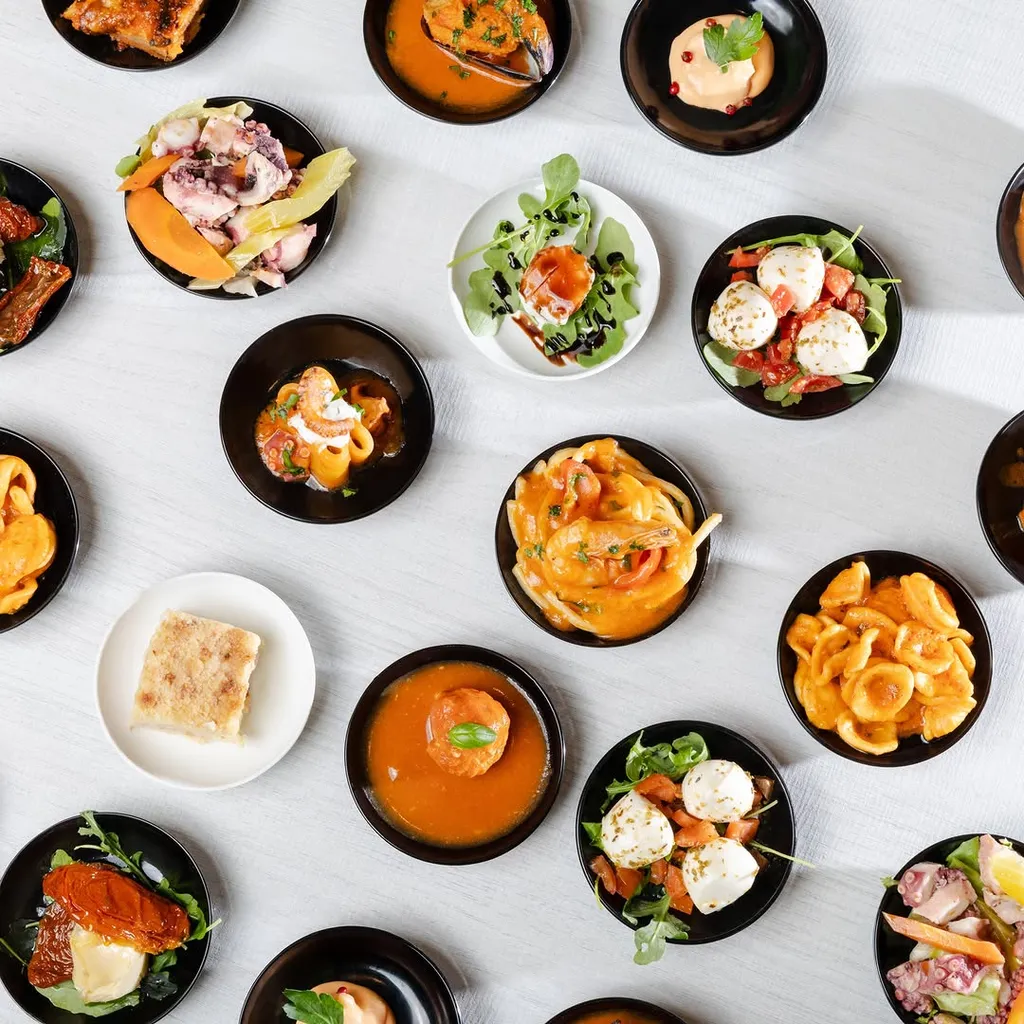 An overhead shot displaying a variety of small, individual portions of food in black and white bowls, arranged neatly on a light-colored surface, showcasing different dishes.