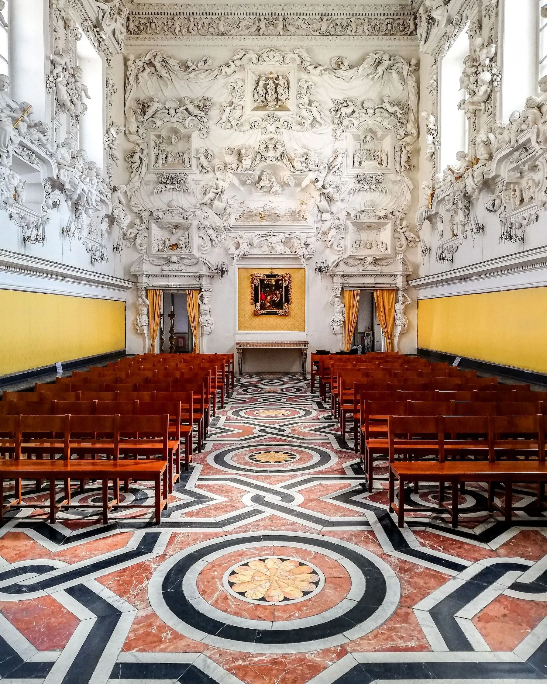 An interior shot of an ornate historical hall, featuring intricate white stucco relief work on the upper walls and ceiling, contrasting with yellow lower walls. The room is furnished with rows of wooden chairs arranged symmetrically, leading towards a painting on the far wall. The floor boasts a striking geometric pattern in red, black, and white marble. The composition is strong, utilizing leading lines from the floor to guide the eye through the space. The lighting is even and natural, highlighting the detailed craftsmanship without harsh shadows. Overall sharpness and color accuracy are excellent.
