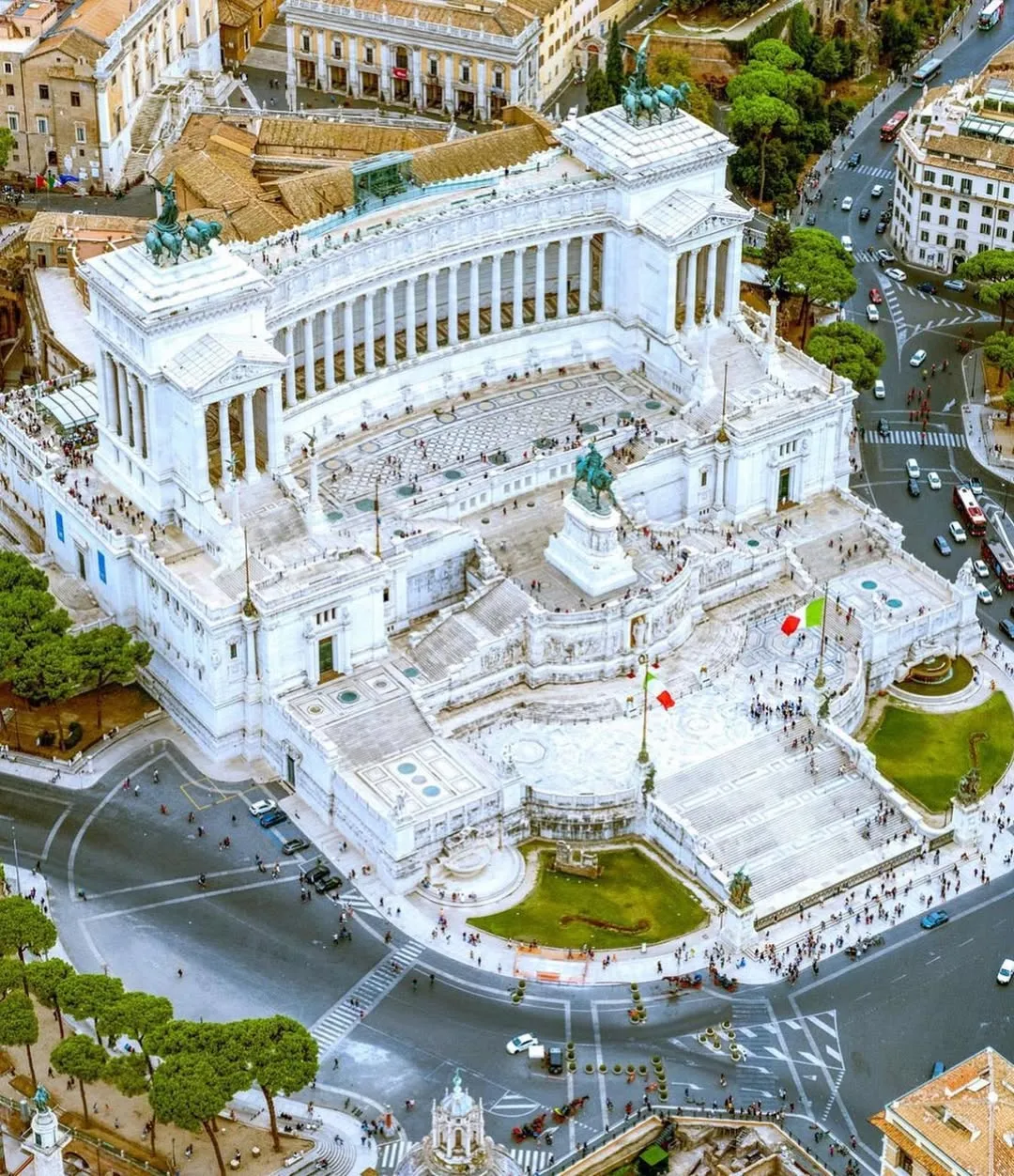 An impressive aerial view of the Altare della Patria (Victor Emmanuel II Monument) in Rome, Italy, showcasing its grandeur and intricate details amidst the city's urban fabric. The white marble structure dominates the frame, surrounded by streets, vehicles, and pedestrians.
