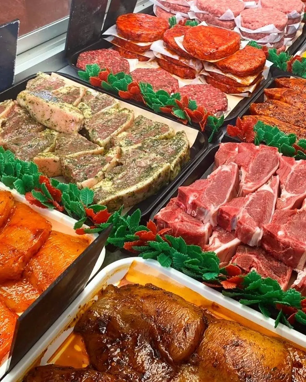 A well-lit and realistic display of various fresh and prepared meat cuts in a retail or butcher shop setting, featuring different types of red meat, some seasoned, neatly arranged in trays with decorative green foliage.