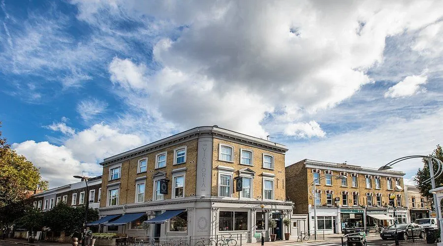An exterior shot of a building named 'The Victoria' under a partly cloudy sky, featuring its architectural details and street-level elements like bicycles.