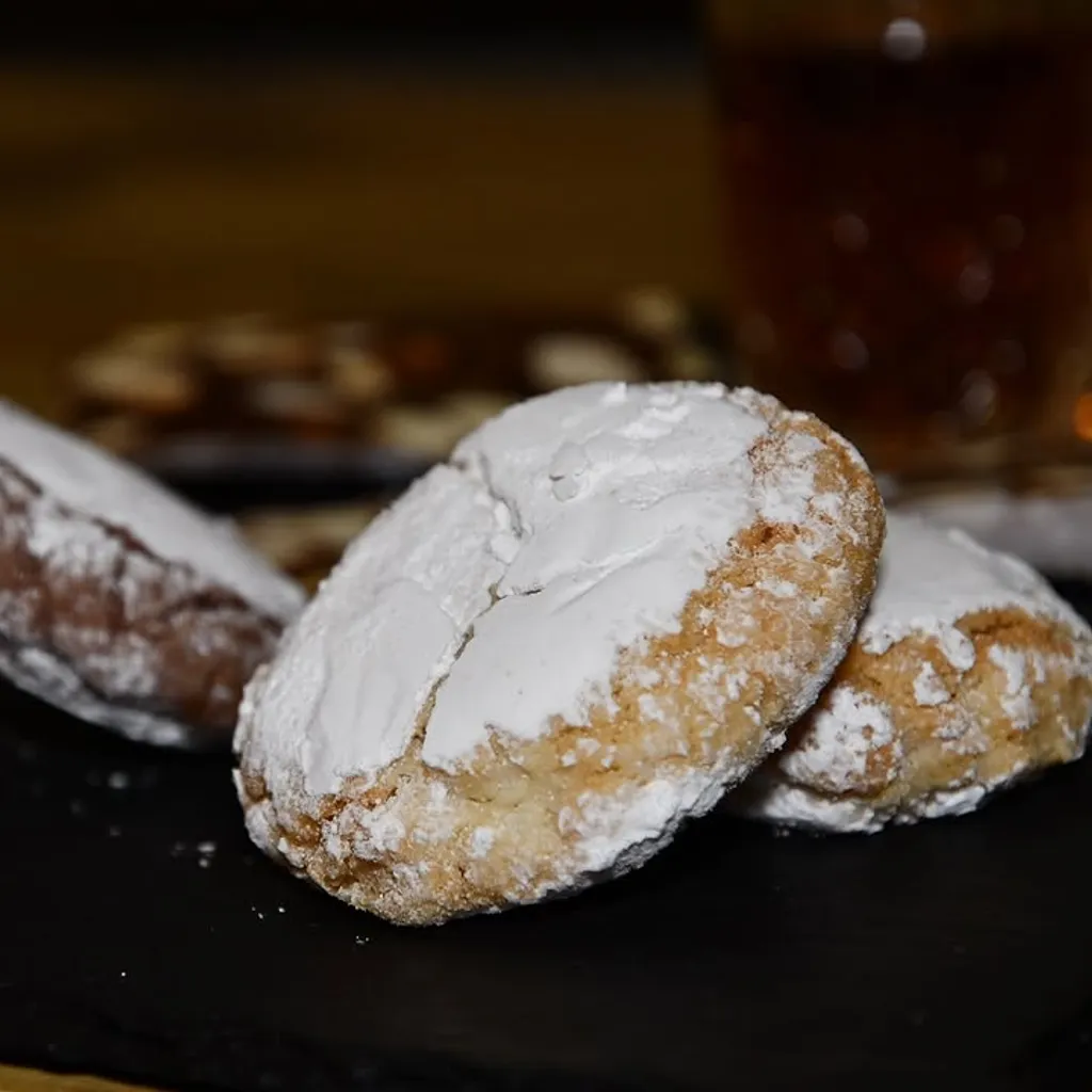 A close-up shot of several sugar-dusted cookies, with one prominently in focus in the foreground on a dark surface. The background shows more blurred cookies.