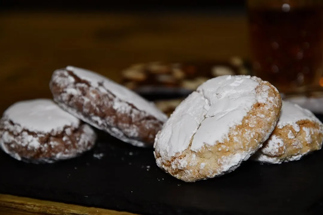 A close-up shot of several sugar-dusted cookies, with one prominently in focus in the foreground on a dark surface. The background shows more blurred cookies.