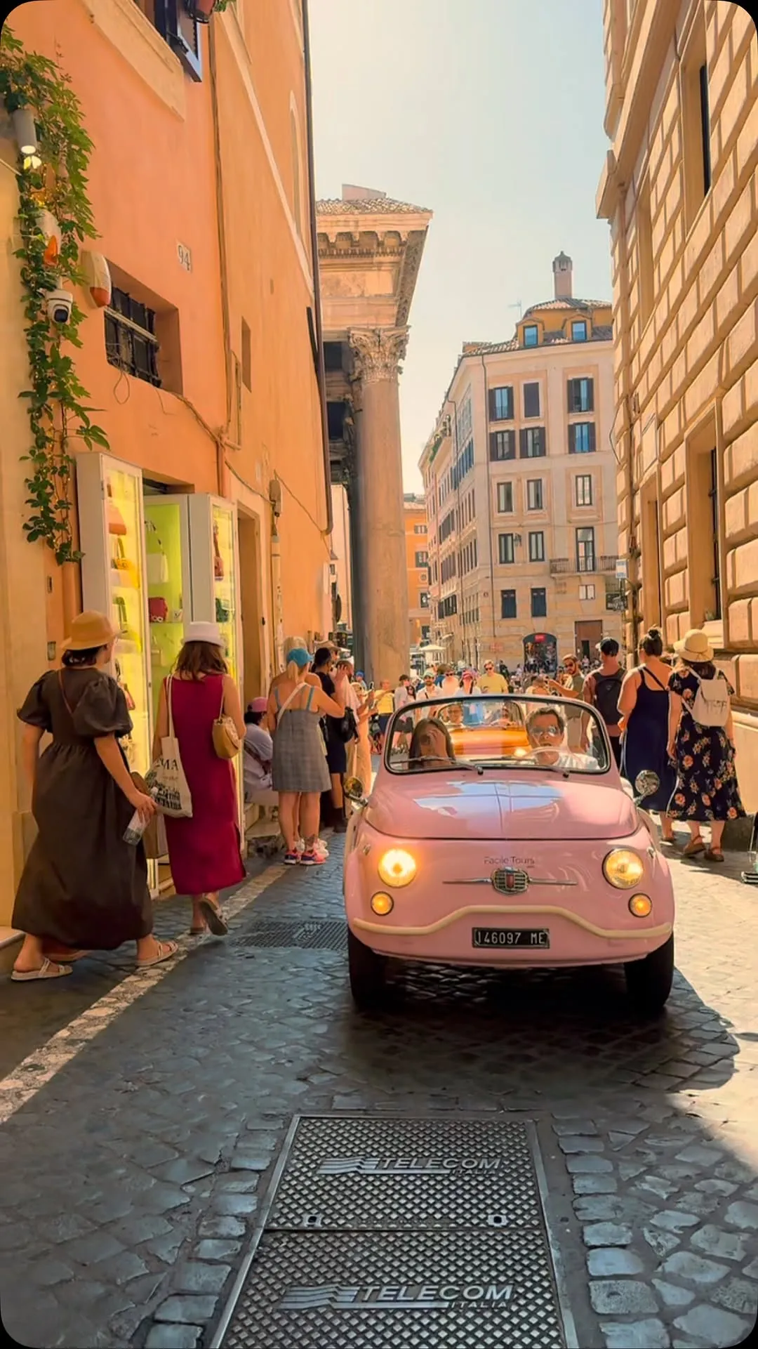 A vibrant street scene in a European city, featuring a pink vintage Fiat 500 driving down a narrow, sunlit cobbled street, surrounded by pedestrians and historic buildings.