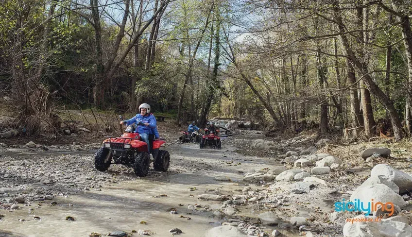 An image featuring three people riding red ATVs through a rocky, muddy riverbed in a wooded area. The lead rider is in the foreground, with two other riders further back creating a sense of depth. The surrounding trees show early spring foliage.