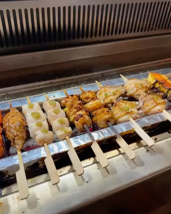 An appealing close-up shot of various skewers cooking on a grill. The composition effectively showcases the different types of food, from chicken to what appears to be scallops or fish cakes, all visibly cooking over heat.