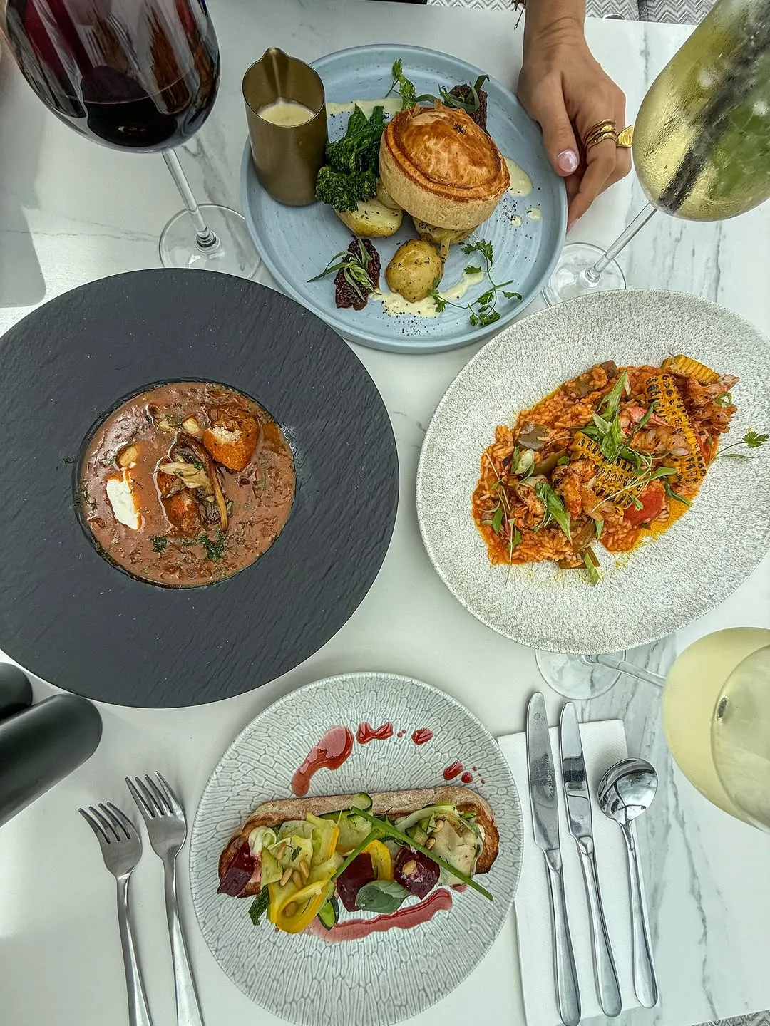 An overhead shot of a dining table showcasing multiple professionally plated dishes, including a pie, a risotto-like dish, and a soup, alongside beverages and cutlery. The image is well-composed with excellent lighting, sharp focus on all key food elements, and accurate, vibrant colors, creating an appealing and photo-realistic presentation of a gourmet meal.