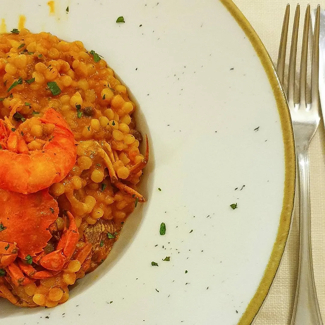 A close-up shot of a seafood pasta dish, possibly Fregola or similar small pasta, featuring shrimp and crab, served on a white plate with cutlery visible on the side.