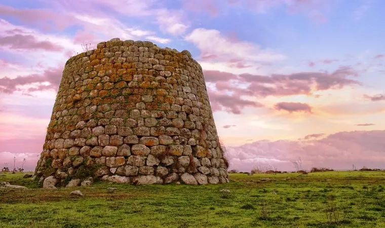An ancient stone structure, likely a Nuraghe, stands prominently on a grassy field against a vibrant sunset sky. The composition is strong, placing the historical building off-center, allowing the dramatic sky to fill the background. The warm light of the setting sun illuminates the rough texture of the stone, and the overall scene evokes a sense of timelessness.