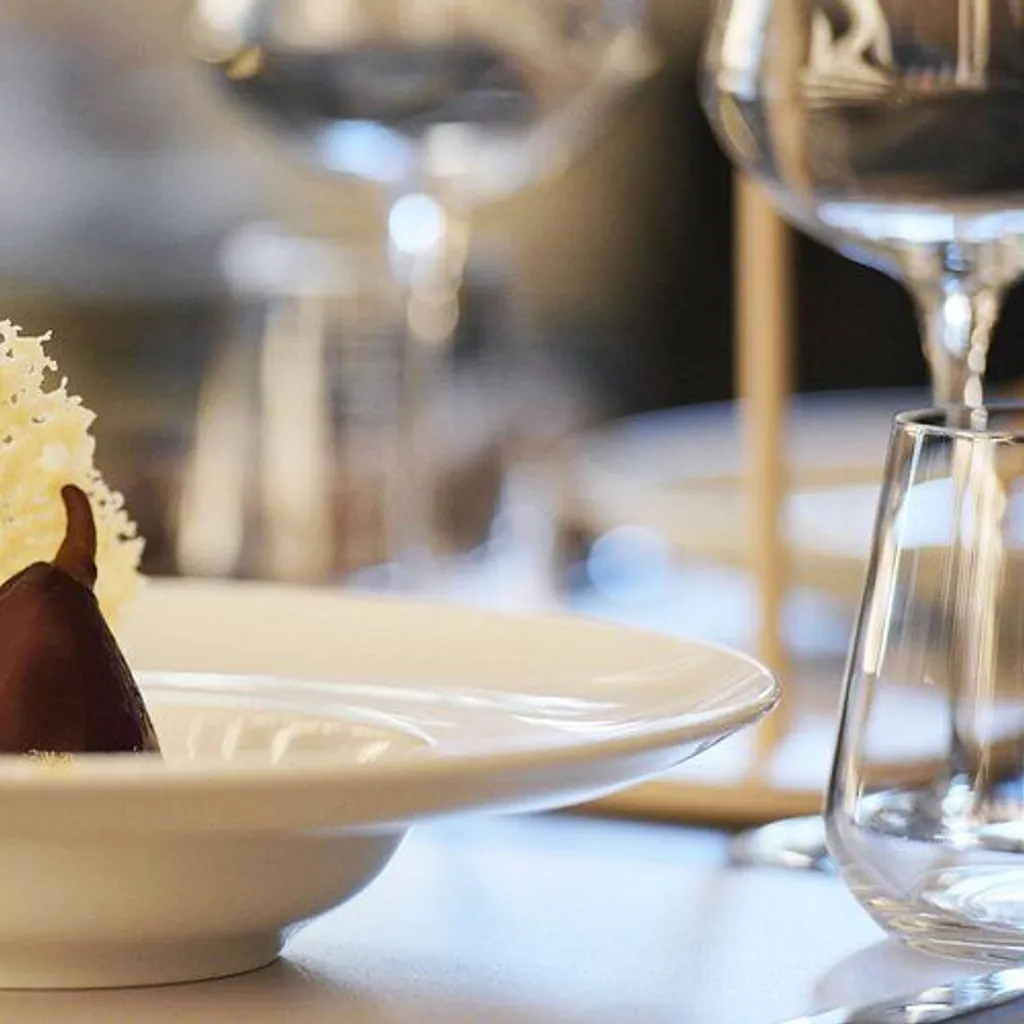 The image features an elegantly plated dessert, appearing to be a chocolate-covered pear with a decorative sugar or cheese crisp. It is presented in a white bowl. The background is softly blurred, showing hints of a glass and other table elements.