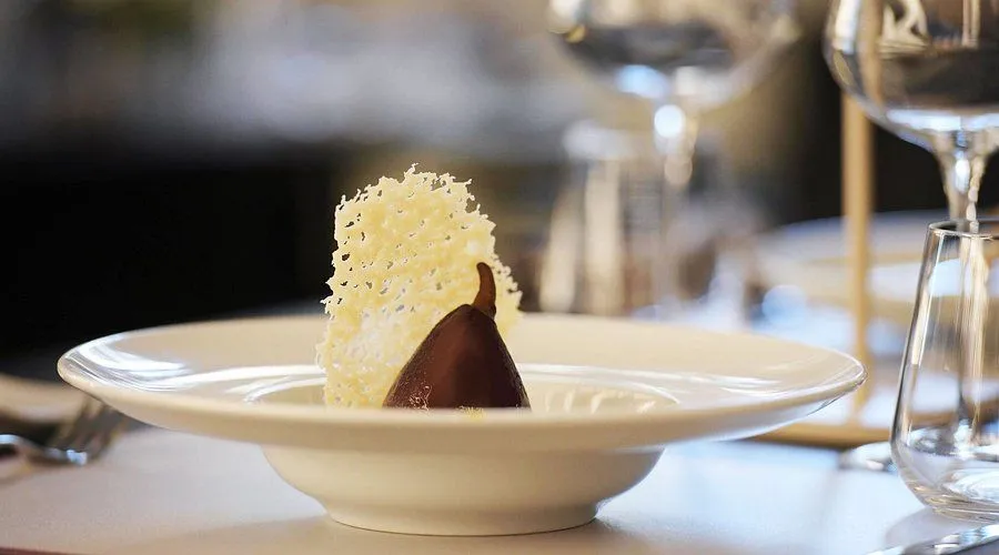The image features an elegantly plated dessert, appearing to be a chocolate-covered pear with a decorative sugar or cheese crisp. It is presented in a white bowl. The background is softly blurred, showing hints of a glass and other table elements.