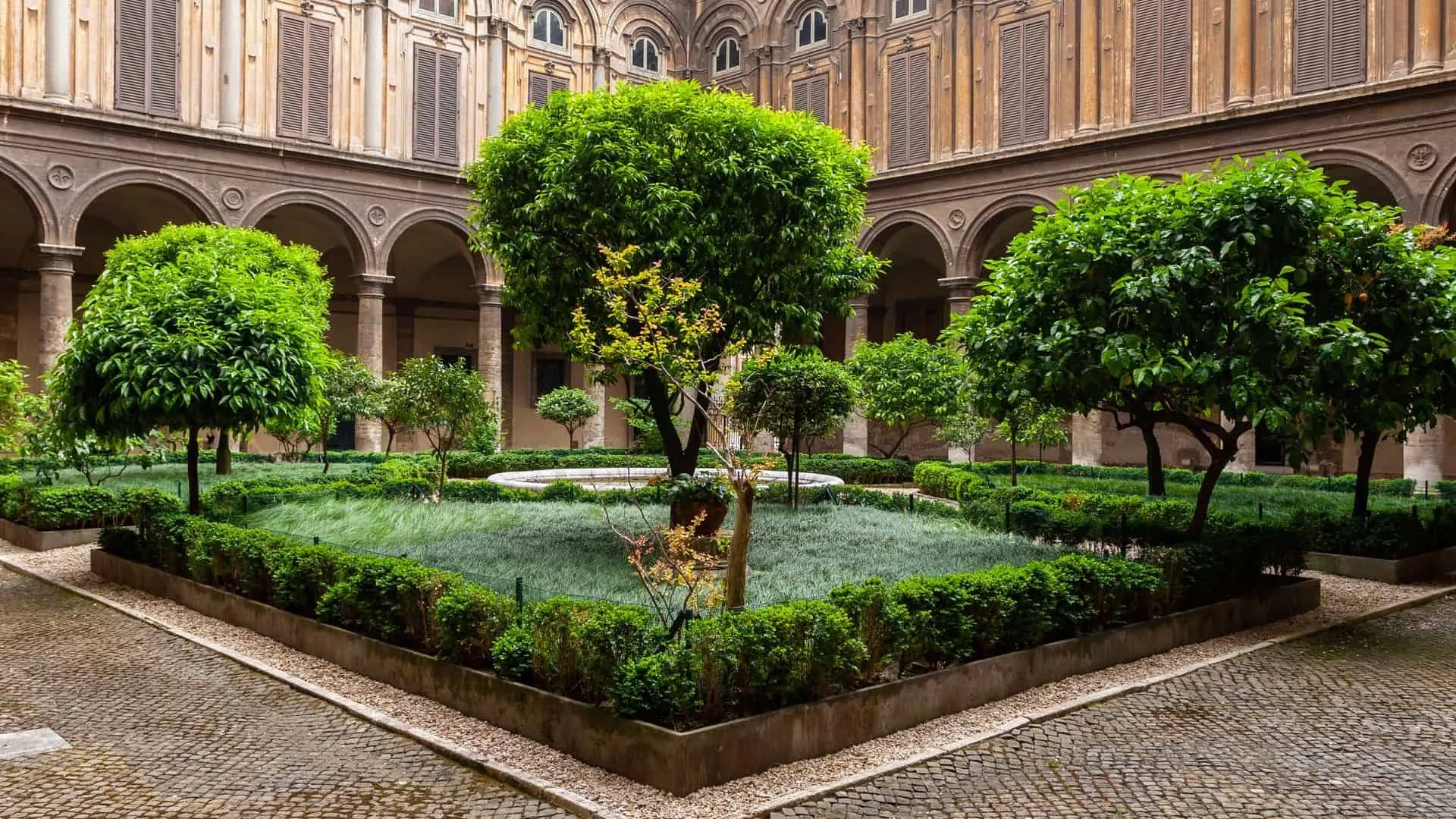 An exquisite view of a meticulously maintained garden courtyard, enclosed by a grand, historic building. The lush greenery, including prominent trees and manicured hedges, contrasts beautifully with the stone architecture. The image showcases architectural details and a serene outdoor space.