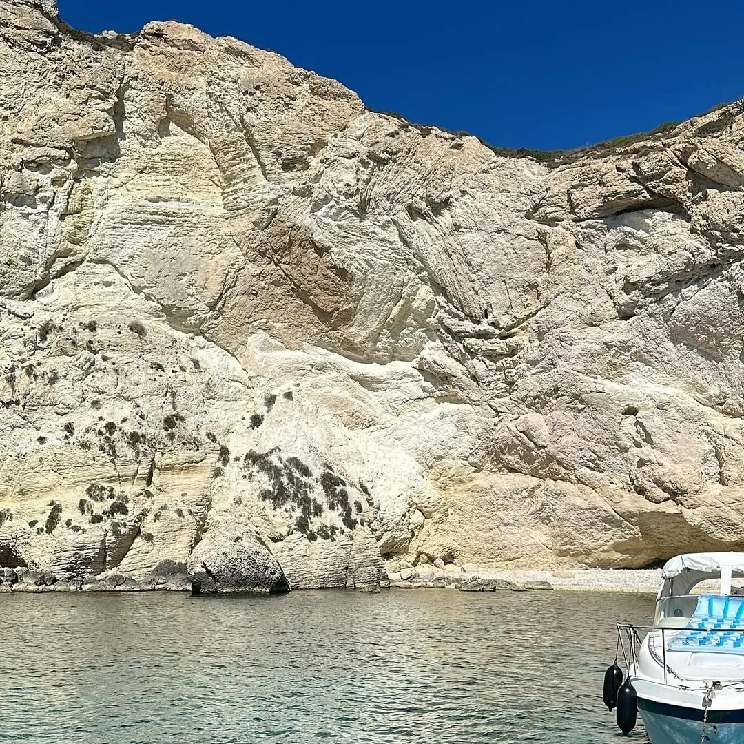 An image featuring a large, light-colored cliff face rising from clear blue-green water under a vibrant blue sky, with a white boat partially visible in the lower right corner.