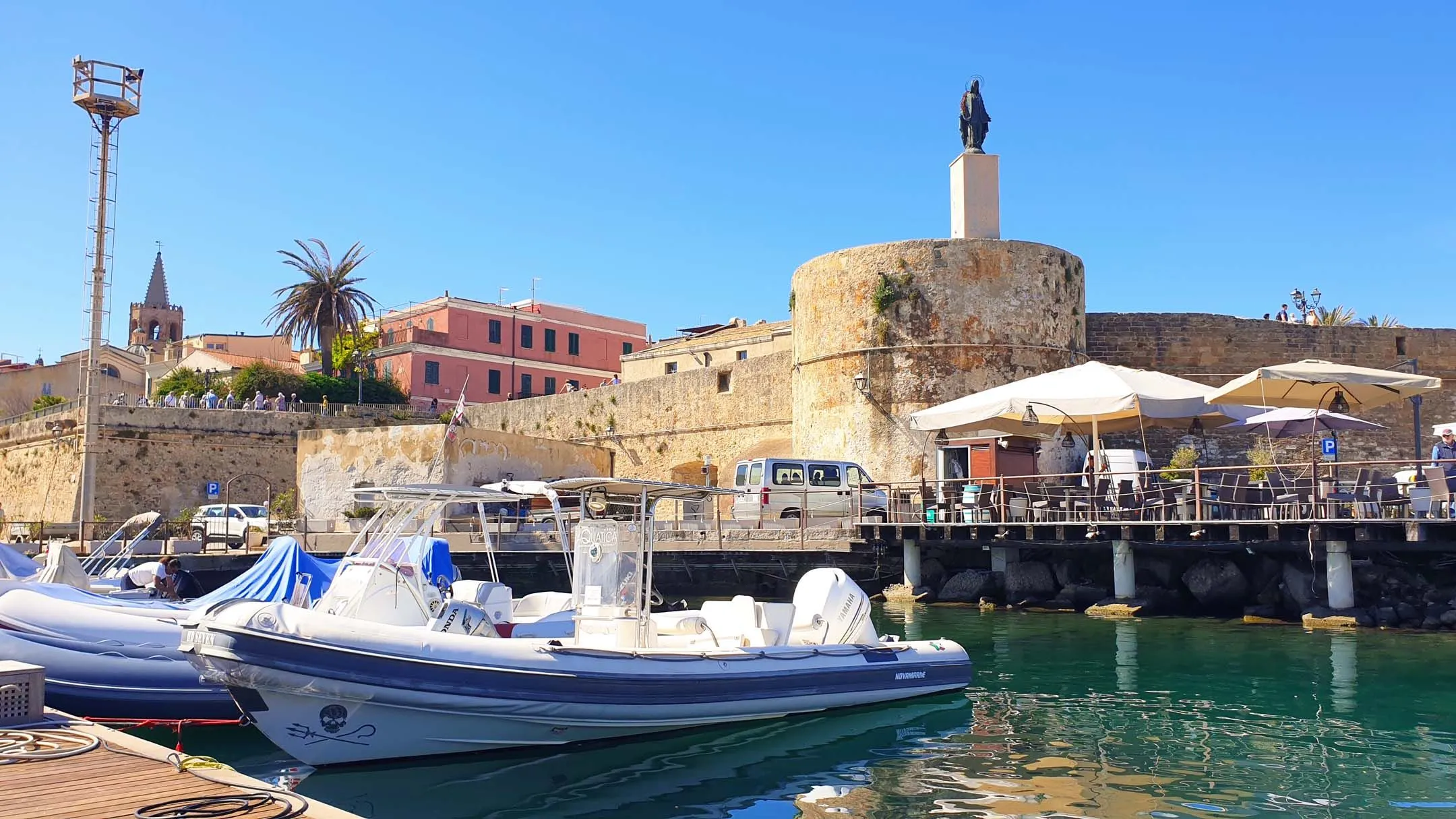 An image of a harbor with a boat in the foreground, a historic stone fort with a statue on top, and buildings in the background under a clear blue sky.