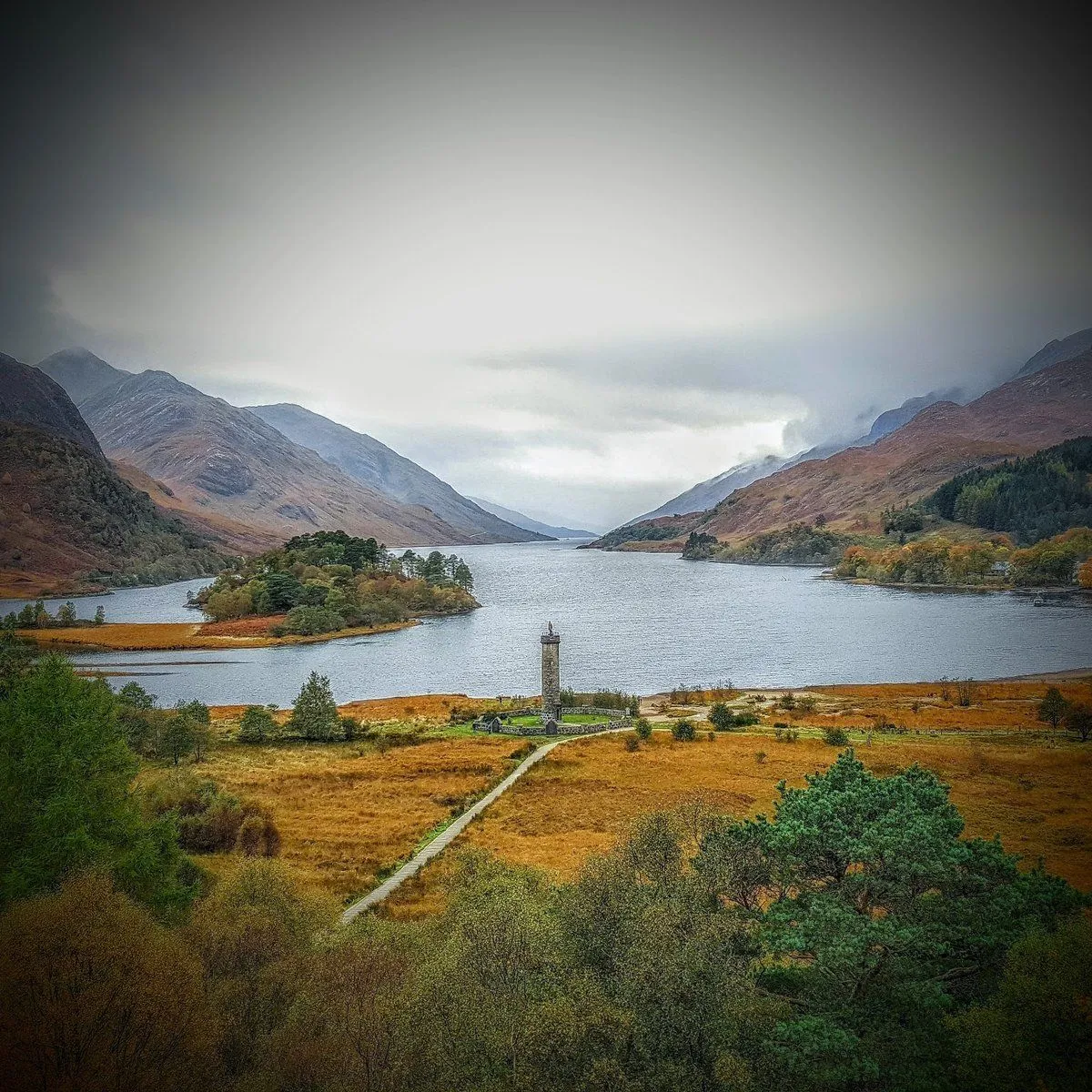 A scenic landscape photograph of a loch surrounded by mountains under an overcast sky, featuring a monument and a path leading towards it.