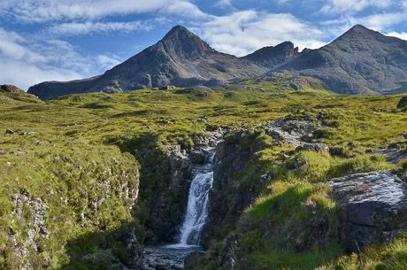 A breathtaking natural landscape featuring a dynamic waterfall cascading over rocky terrain, leading the eye towards a prominent, rugged mountain peak under a vibrant blue sky with scattered clouds. Lush green grass covers the hillsides.