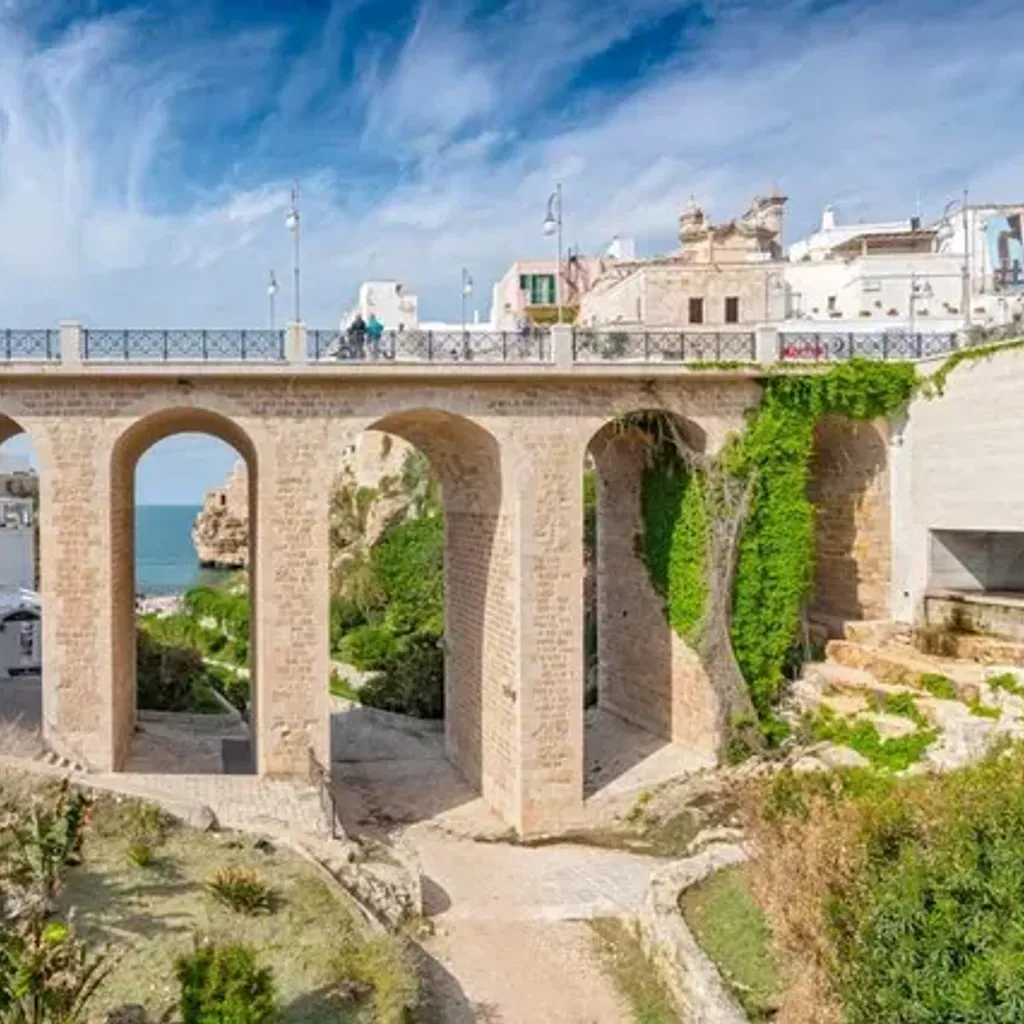 A picturesque view of an ancient stone bridge with multiple arches spanning a gorge, revealing the blue sea and a rocky coastline in the background. The bridge is well-preserved with classic architecture, and lush green vegetation adorns the cliffs.