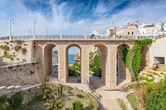 A picturesque view of an ancient stone bridge with multiple arches spanning a gorge, revealing the blue sea and a rocky coastline in the background. The bridge is well-preserved with classic architecture, and lush green vegetation adorns the cliffs.