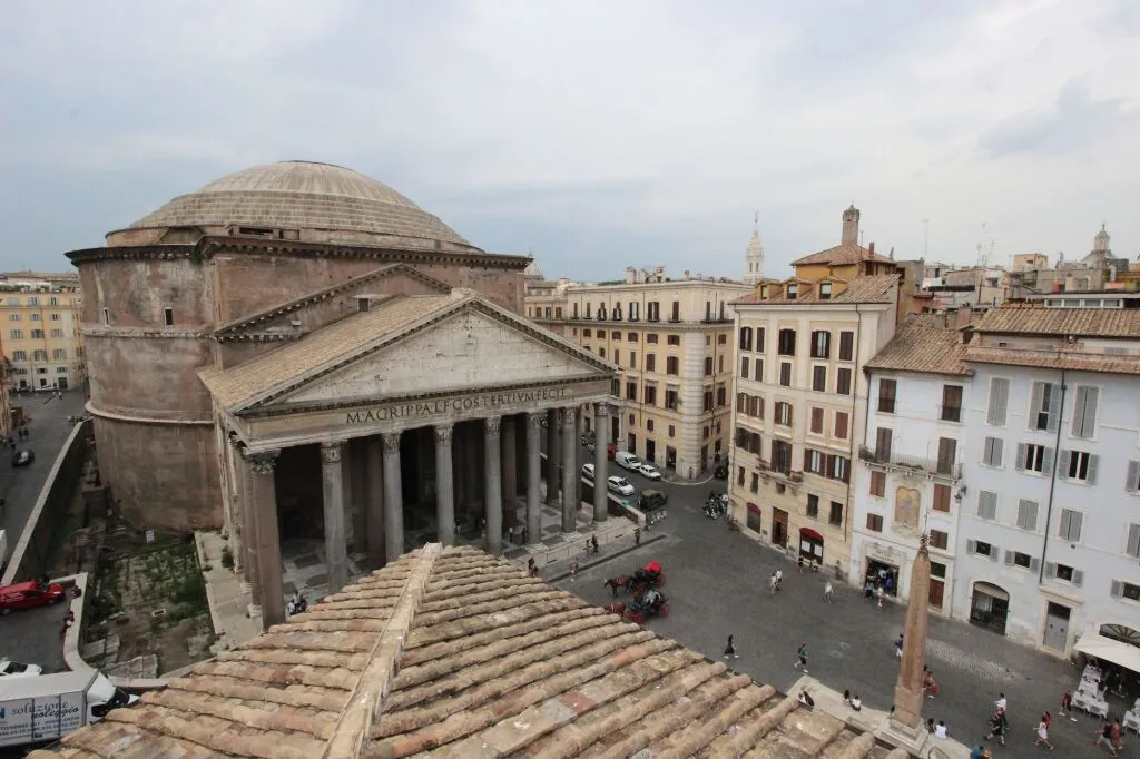 An elevated view of the Pantheon and the surrounding piazza in Rome, Italy, captured on what appears to be an overcast day. The image features the iconic Roman temple with its large columns and dome, set against a backdrop of historic city buildings and a glimpse of an obelisk in the square below.