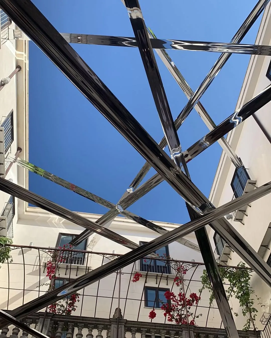A view looking upwards towards a clear blue sky, framed by white buildings, with a prominent, intersecting metal structure in the foreground that creates strong geometric patterns and reflections.