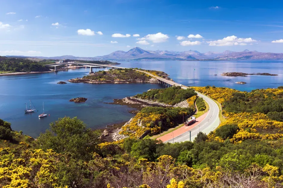 An outstanding panoramic landscape photograph showcasing a bridge spanning across a body of water with lush green and yellow vegetation in the foreground and majestic mountains in the background under a clear blue sky. The image exhibits excellent composition and vibrant, natural colors.