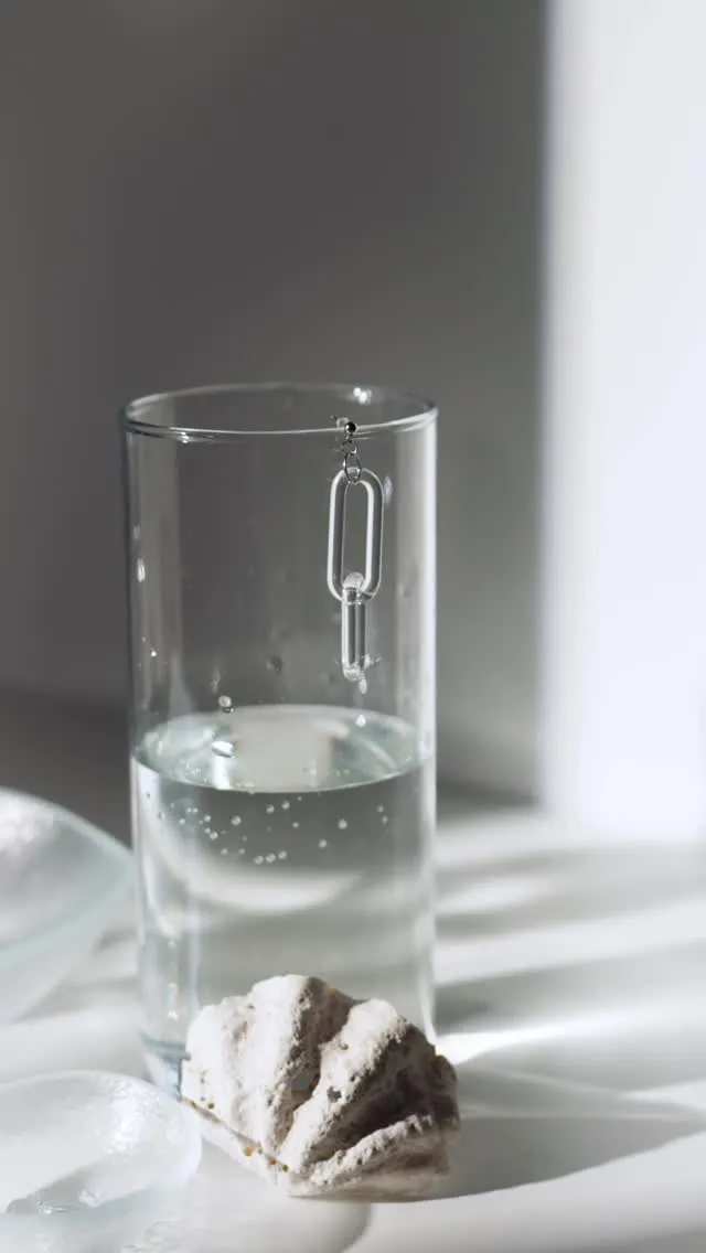 The image features a transparent chain-link earring suspended over a glass of water. The composition is clean and minimalist, focusing on the intricate design of the earring and its clear material.