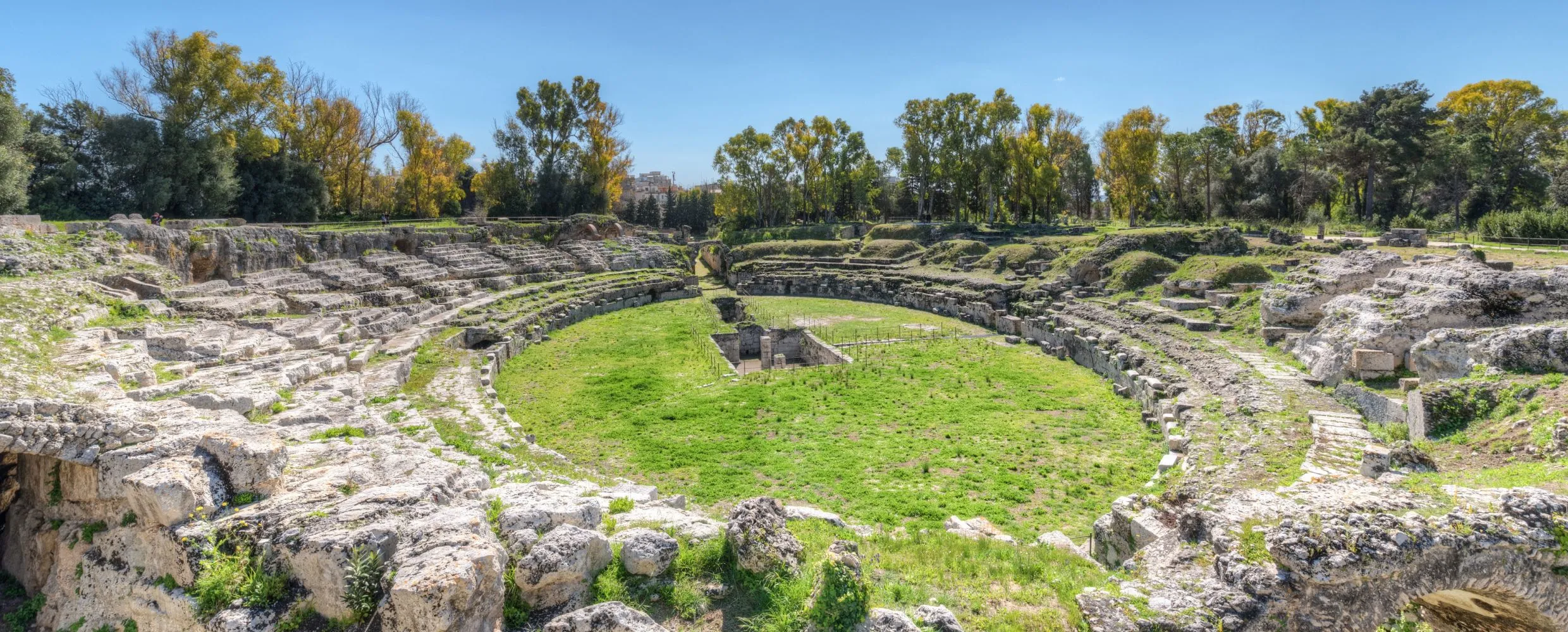 An outdoor shot of an ancient amphitheater, featuring stone seating and a grassy arena under a clear blue sky. The image has good natural lighting, vibrant colors, and sharp details.