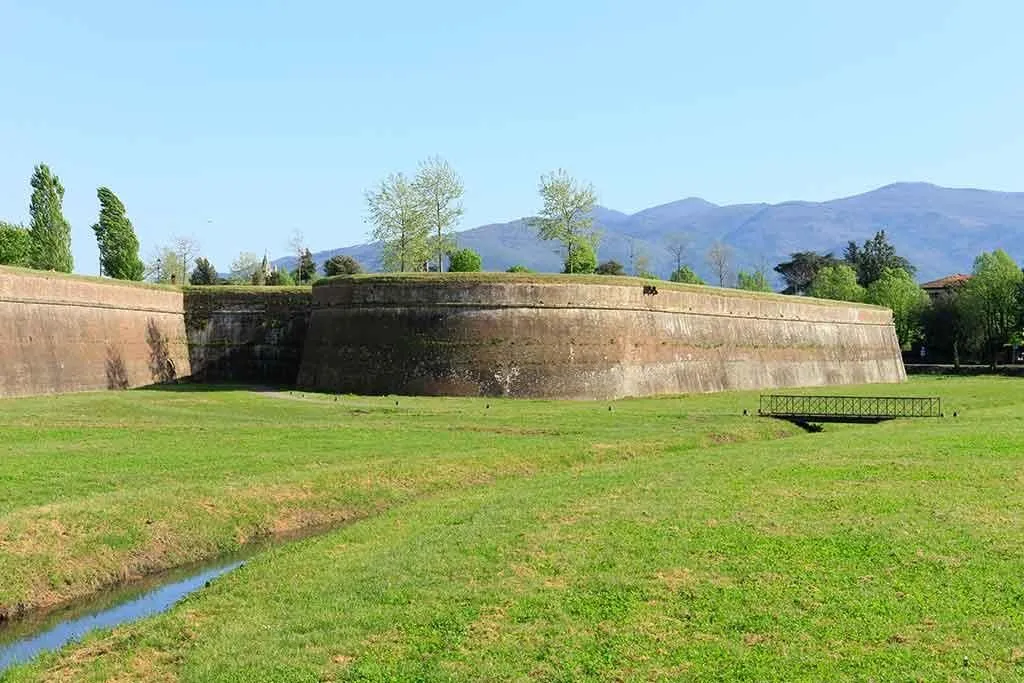 A well-preserved ancient city wall with a moat and grassy foreground under a clear blue sky, with distant mountains.