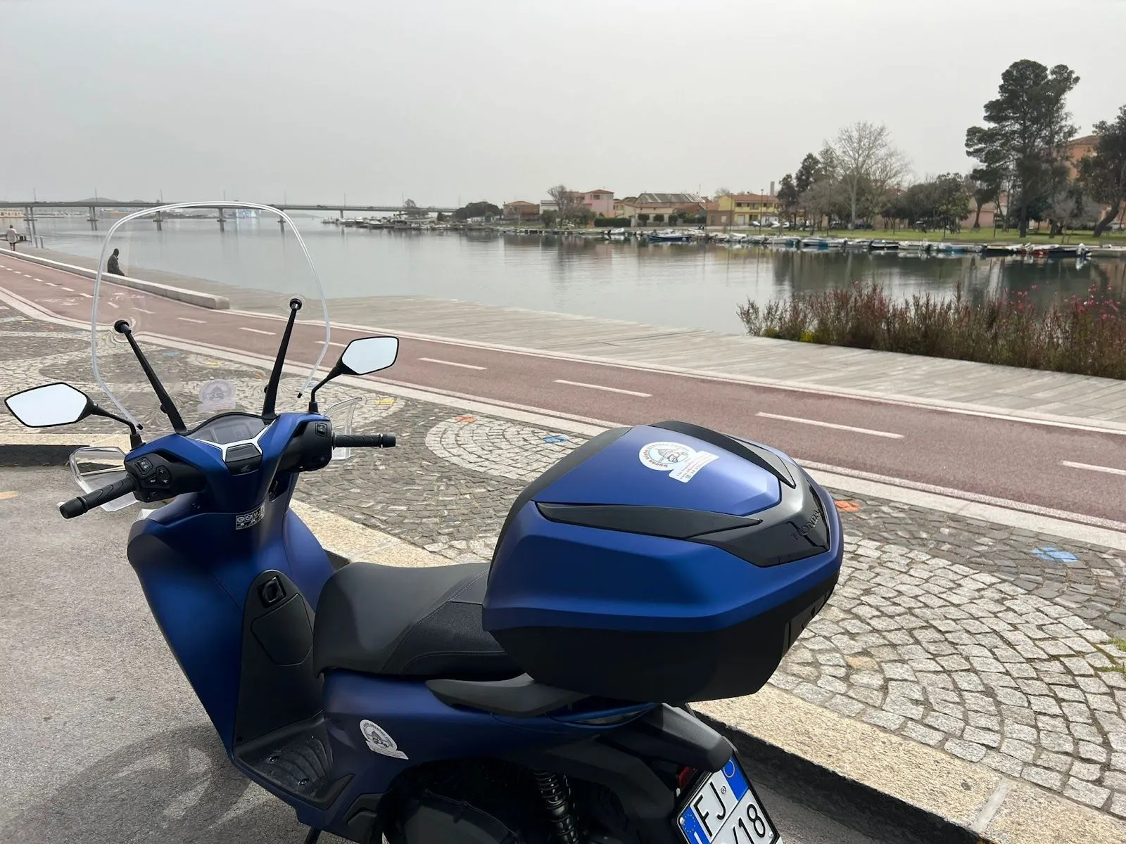 A blue scooter is parked on a cobblestone path next to a wide river or canal, with a bridge and buildings visible in the distance under an overcast sky.