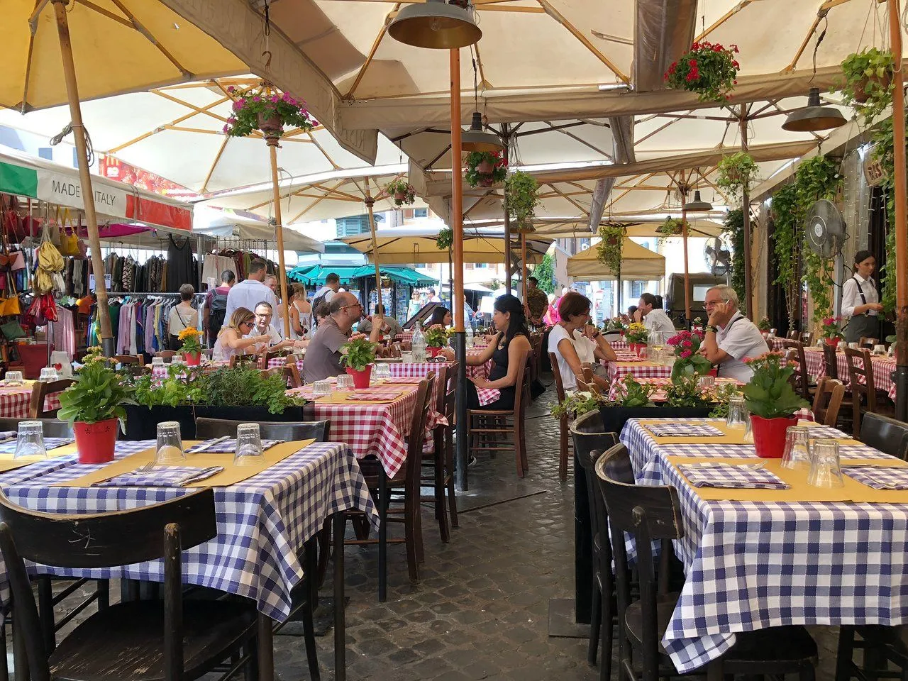 The image successfully conveys the bustling atmosphere of an outdoor dining area, likely within a market setting. It benefits from natural, even lighting and accurate colors, enhancing its photo-realistic quality. The sharpness is generally good for the key elements in the mid-ground, although the composition is somewhat casual, with elements like foreground tables being partially cut off and a busy background. Despite the active environment, the scene is well-captured in an editorial style.