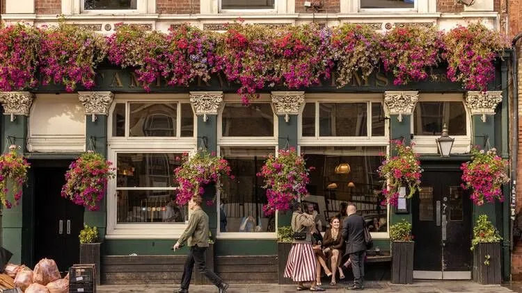 A vibrant and well-composed street scene featuring the facade of a building adorned with abundant pink and purple flowers. The image is sharp, photo-realistic, and exhibits good color accuracy. People are visible on the sidewalk, adding to the urban feel of the shot. Key elements are in focus, creating a clear and engaging visual.