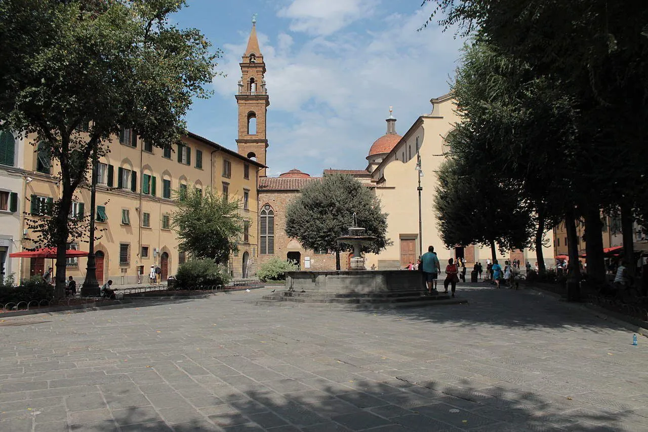 An inviting editorial-style image capturing a sunny piazza in Italy, featuring a historical fountain, lush green trees, and an impressive bell tower and church in the background. The natural light and color accuracy create a realistic and pleasant scene.