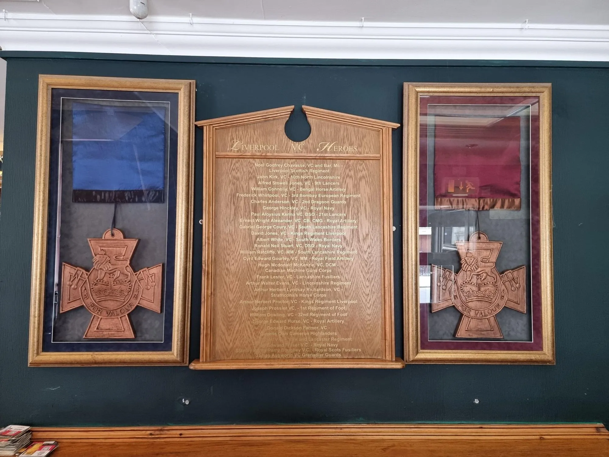 An image showcasing a historical display featuring a wooden plaque listing "Liverpool VC Heroes" flanked by two framed medals and fabric, set against a dark wall.
