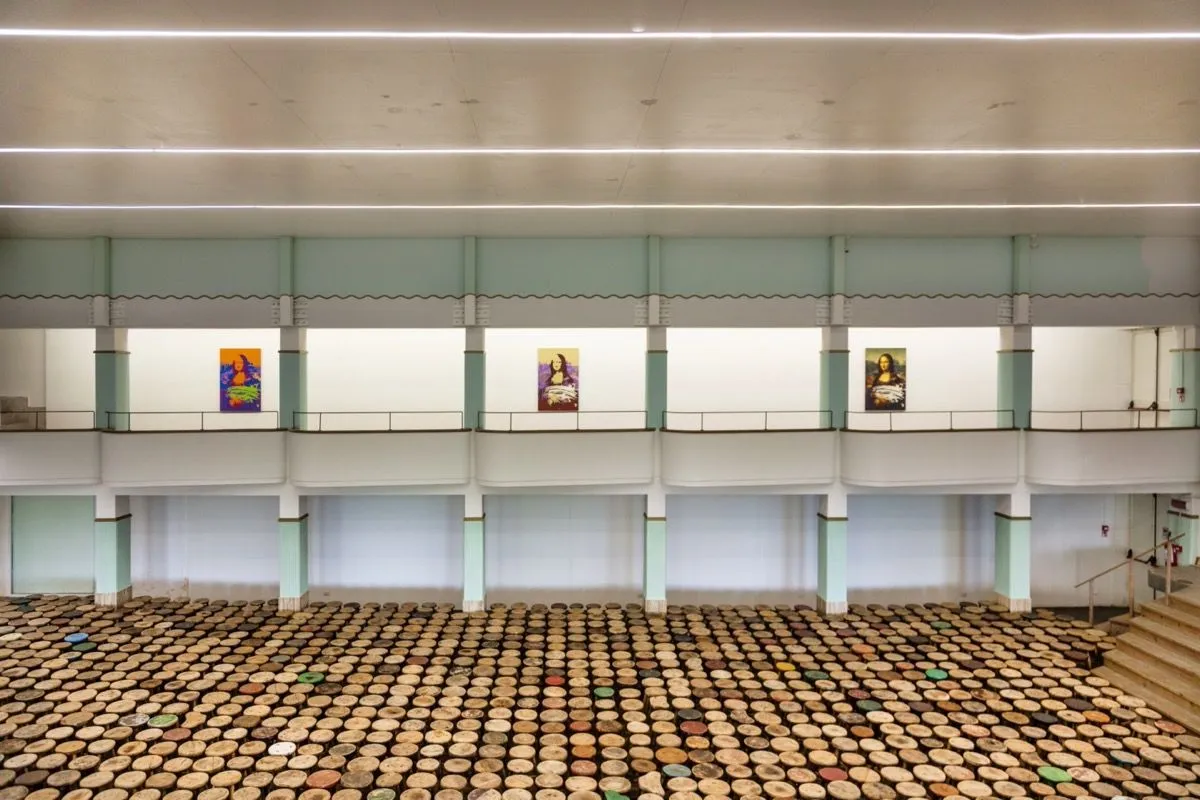 An indoor wide shot of an art exhibition space featuring multiple framed artworks resembling Mona Lisa displayed on an upper wall, and a large floor installation consisting of numerous circular, flat objects, some painted with various colors.