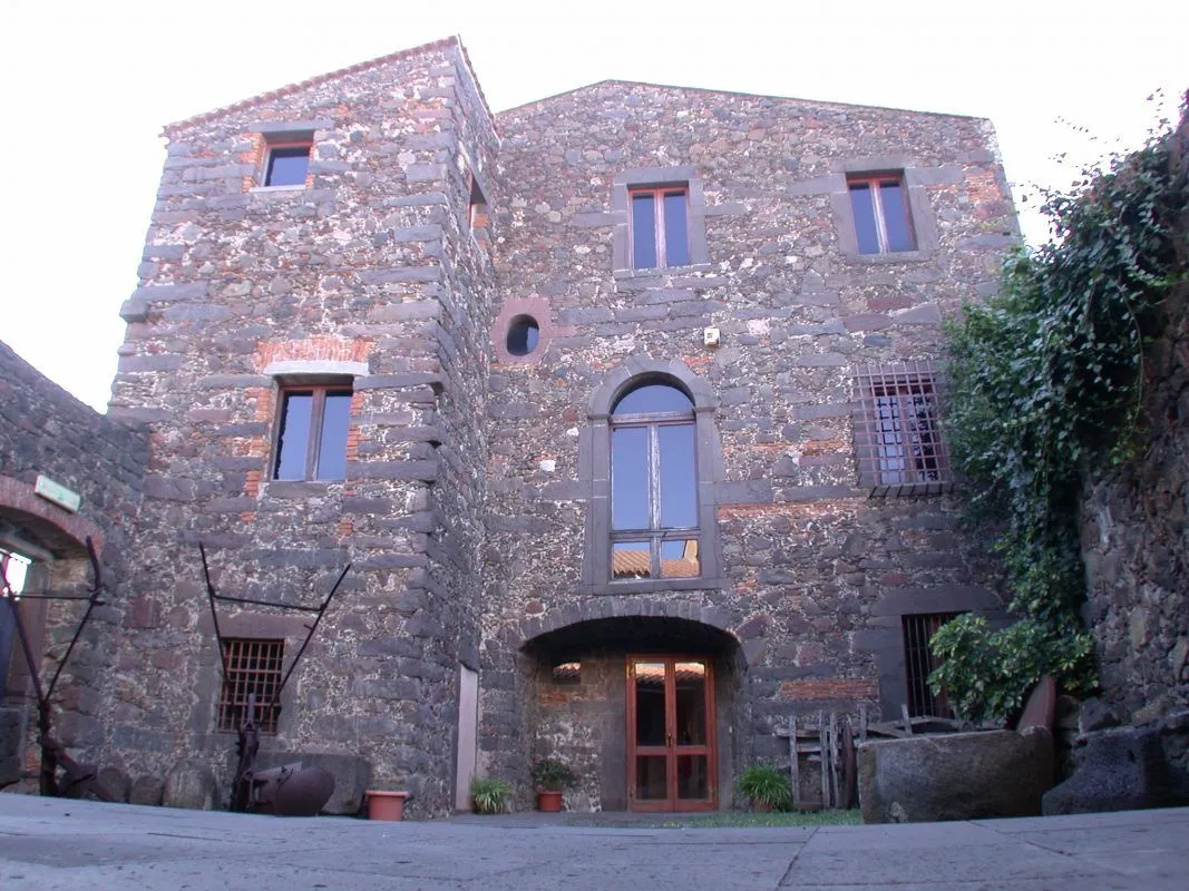 An exterior view of an old multi-story building constructed from dark grey stone, featuring various windows, some with wooden frames and one with iron bars. A large arched entryway leads to a wooden door. The building has multiple rooflines and appears historic, set in a rustic environment.