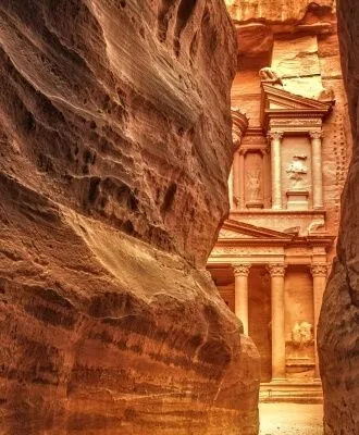 An iconic view of Al-Khazneh (The Treasury) in Petra, Jordan, seen through the narrow Siq canyon. The ancient rock-cut facade is framed by the natural sandstone walls, showcasing its intricate details and the warm, earthy tones of the surrounding landscape.