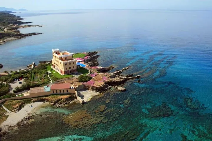 The image showcases a well-composed scene with a striking building on a rocky coastline, featuring crystal-clear blue waters. The natural lighting is excellent, illuminating the scene without harsh shadows or blown-out highlights. The colors are vibrant and accurate, particularly the varying shades of blue in the sea. The image is generally sharp and highly photo-realistic, with key elements like the building and foreground rocks in good focus. Minor improvements could be made to the crispness of very distant elements.