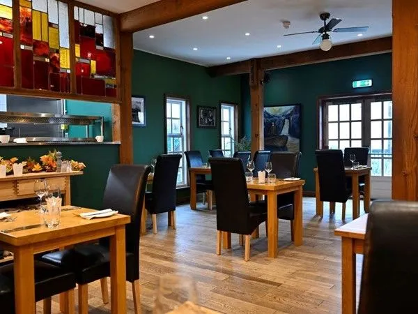 An editorial quality image of a restaurant interior. The composition effectively showcases the dining area with multiple tables and chairs, accented by distinctive green walls, wooden beams, and a stained glass feature. The lighting is generally good, creating a warm and inviting atmosphere. The image is sharp and color-accurate, contributing to its photo-realistic quality. All key elements are in focus, providing a clear view of the space.