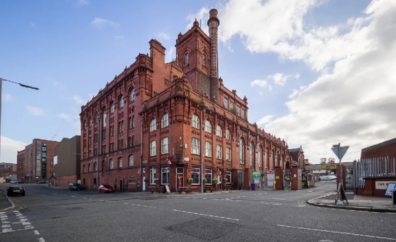 An exterior shot of a large, old red-brick building with intricate architectural details, a prominent chimney, and multiple windows, situated on an urban street corner under a blue sky with clouds.